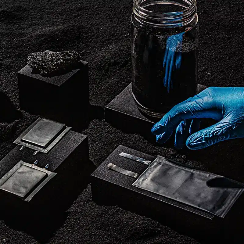 A hand wearing a blue lab glove reaches over battery cells and materials