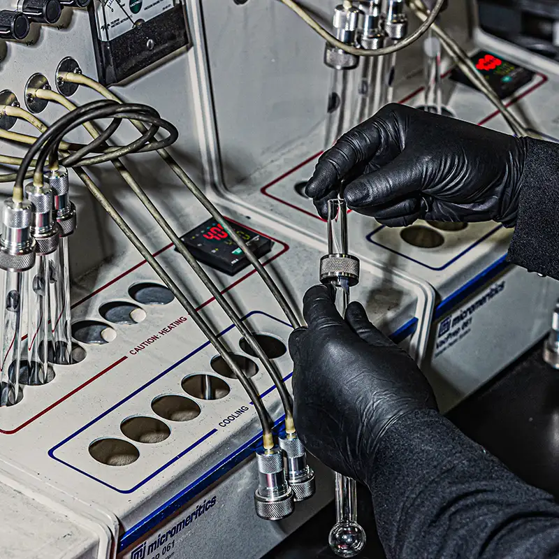 A close-up of a technician's hands in black lab gloves preparing material for BET surface area measurement